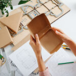 Female box maker working behind a table in a private workshop, folding, shaping a box, tools scattered across. Rulers, pencils and special board with a cutting form.