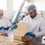 Young Caucasian employee in sterile uniform packing goods in boxes. In background supervisor holding tablet and counting boxes.