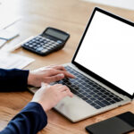 Businesswomen hand on mockup laptop keyboard with blank screen on the desk, White screen laptop and smartphone with a calculator on the table, girls using blank screen laptop with a phone and office equipment on the desk. Close - up shot.